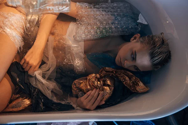 Top view of young woman lying in bathtub with trash and looking away, environmental pollution concept — Stock Photo