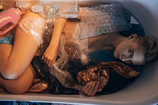 Вид сверху на молодую женщину, лежащую в ванной с мусором, концепция загрязнения окружающей среды — стоковое фото