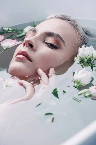 Schöne Frau, die im klaren Wasser mit Blumen liegt und in die Kamera schaut — Stockfoto