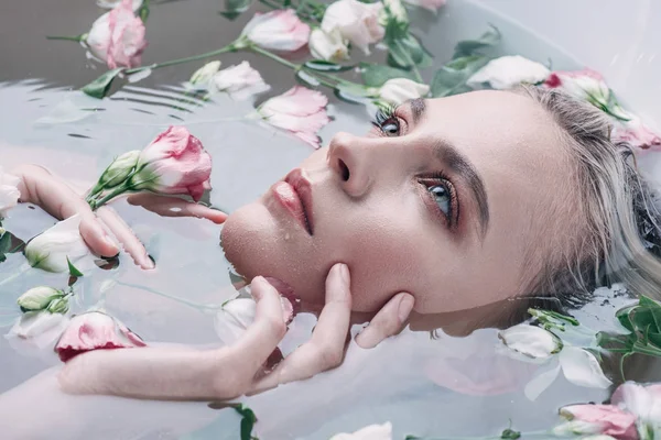 Schöne Frau liegt im klaren Wasser mit Blumen in weißer Badewanne und schaut weg — Stockfoto