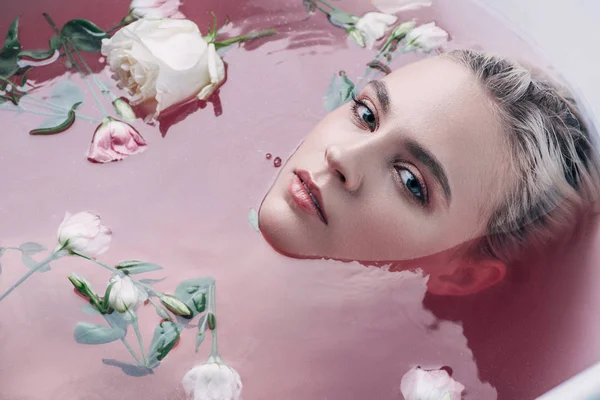 Bella giovane donna sdraiata nella vasca da bagno con acqua rosa colorata e fiori e guardando la fotocamera — Foto stock