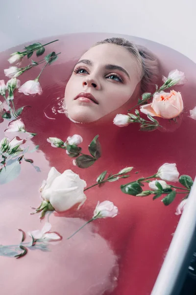 Schöne junge Frau liegt in der Badewanne mit farbigem rosa Wasser und Blumen und schaut weg — Stockfoto
