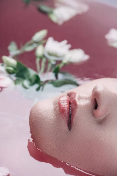 Vue rapprochée de belles lèvres de jeune femme dans de l'eau rose colorée avec des fleurs — Photo de stock