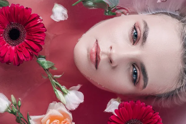 Draufsicht der schönen jungen Frau in der Badewanne liegend mit farbigem rosa Wasser, Gerberas und Rosen — Stockfoto