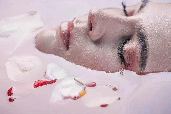 Vue rapprochée de belle femme aux yeux fermés dans de l'eau rose avec des pétales de rose — Photo de stock