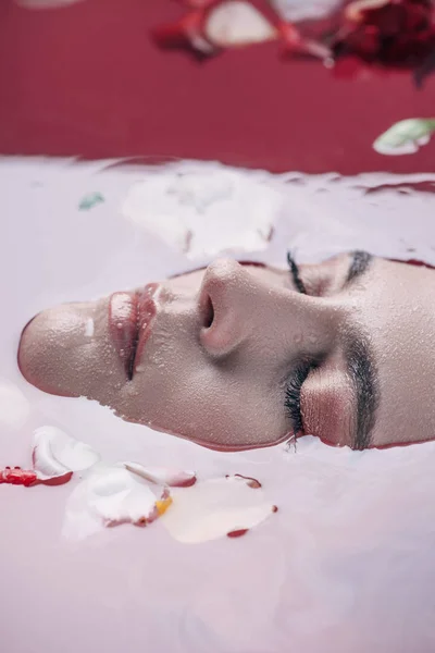 Nahaufnahme der schönen Frau mit geschlossenen Augen in rosa Wasser mit Rosenblättern — Stockfoto