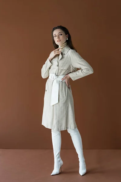 Modelo elegante en gabardina beige de pie con la mano en la cadera aislado en marrón - foto de stock