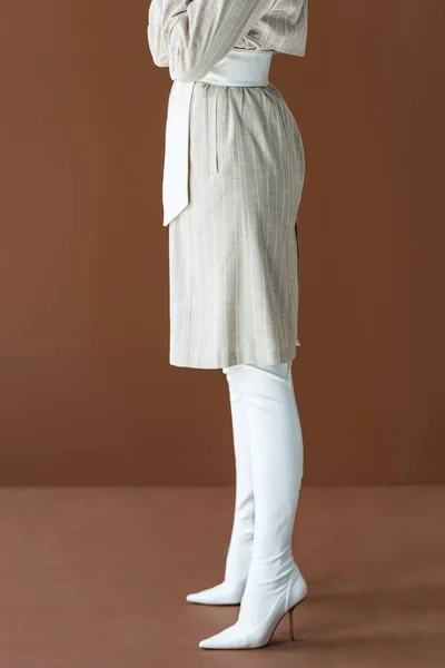 Vista cortada de mulher elegante em casaco de trincheira e botas brancas de pé isolado em marrom — Fotografia de Stock