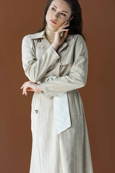 Stylish woman in trendy trench coat isolated on brown, holding hand near face — Stock Photo