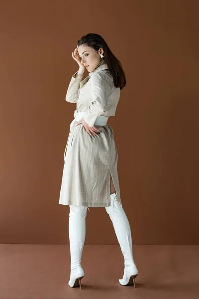 Femme élégante en trench coat beige tendance et bottes blanches isolées sur brun — Photo de stock