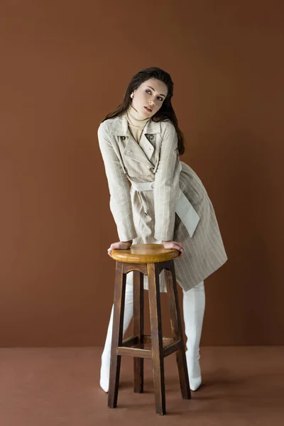 Attrayant adulte femme debout près de chaise isolé sur brun, regardant loin — Photo de stock