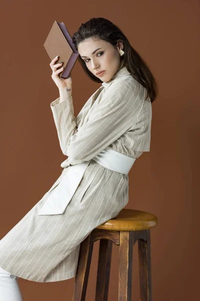 Вид сбоку красивой женщины с книгой в руке, изолированной на коричневом фоне — стоковое фото