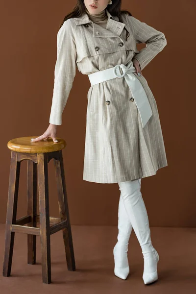 Vista cortada da mulher na moda em casaco de trincheira elegante em pé perto da cadeira isolada no marrom — Fotografia de Stock