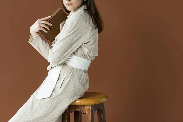 Vista cortada de jovem modelo segurando livro, sentado isolado em marrom — Fotografia de Stock