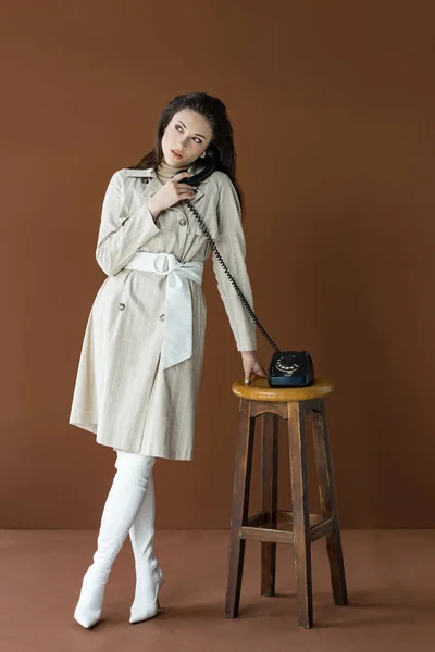 Beautiful model in trench coat talking on telephone, looking away and standing isolated on brown — Stock Photo