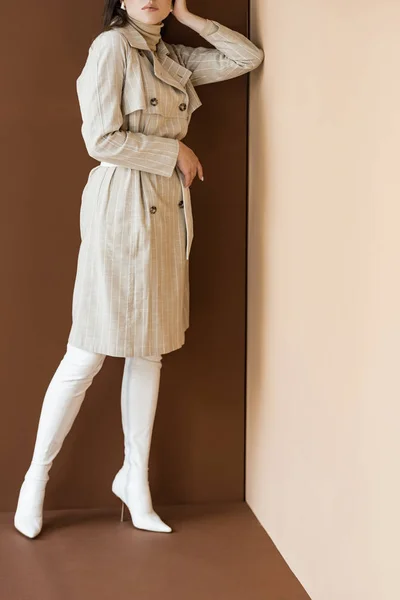 Cropped view of fashionable woman in trench coat and white boots standing on brown background — Stock Photo