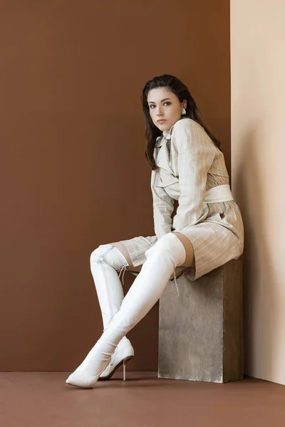 Stylish woman in beige trench coat and white boots sitting on brown background — Stock Photo