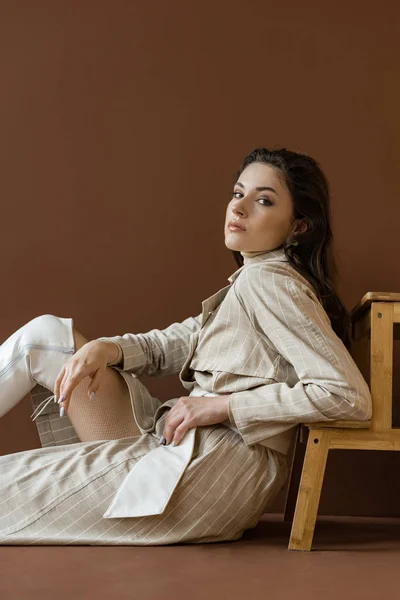 Стильная модель в модном плаще сидит на полу с коричневым фоном, смотрит в камеру — стоковое фото