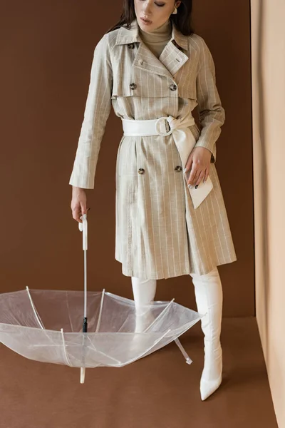 Vista de ángulo alto de la mujer con estilo en la trinchera de moda que sostiene el paraguas, de pie sobre fondo marrón - foto de stock