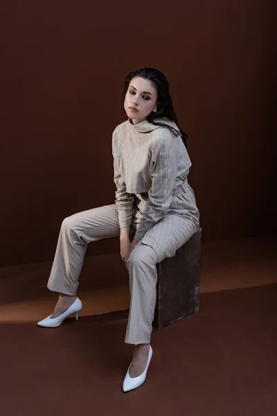 High angle view of stylish woman in trendy wear sitting on box with brown background, looking away — Stock Photo