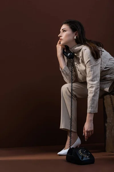 Vista lateral de la mujer con estilo en ropa de moda hablando por teléfono, mirando hacia otro lado y sentado sobre fondo marrón - foto de stock