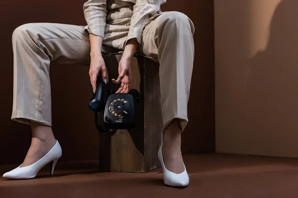 Vista recortada de modelo joven con teléfono retro en manos sentadas sobre fondo marrón - foto de stock