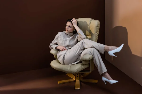 Модная модель сидит на кресле, скрещивает ноги, смотрит в сторону — стоковое фото