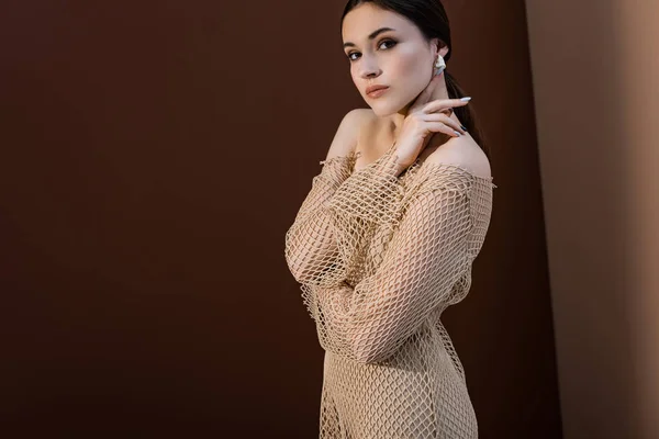 Side view of attractive woman in mesh dress standing on brown background, looking at camera — Stock Photo