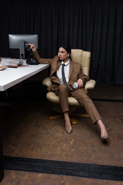 Belle femme d'affaires avec combiné et tasse en papier assis sur un fauteuil dans le bureau, regardant loin — Photo de stock