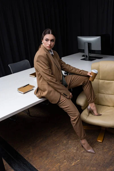 Succès et belle femme d'affaires assise sur la table avec tasse en papier à la main, regardant loin — Photo de stock