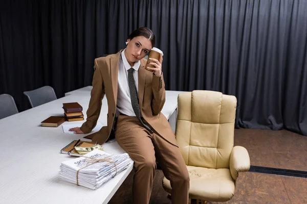 Красивая деловая женщина сидит на столе с бумажной чашкой в руке, глядя на камеру — стоковое фото