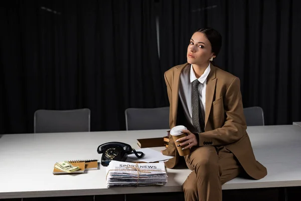 Stylish businesswoman sitting on table in office with paper cup in hand, crossing legs, looking at camera — Stock Photo