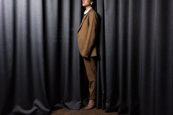 Вид сбоку на модную модель в костюме, стоящую в студии на сером фоне занавеса — стоковое фото