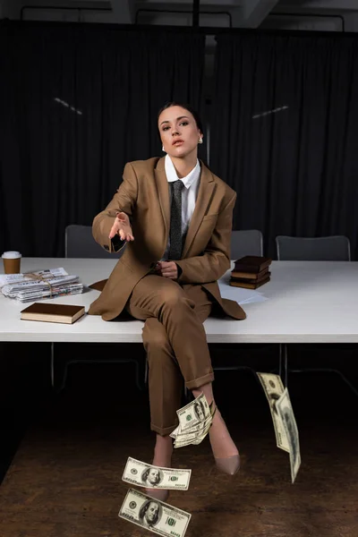 Adult businesswoman sitting on table, crossing legs, looking at camera and throwing money — Stock Photo