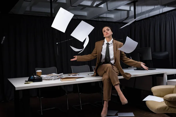 Geschäftsfrau sitzt auf dem Tisch unter fallenden Papierrollen, kreuzt die Beine, blickt in die Kamera — Stockfoto