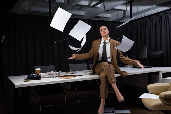 Femme d'affaires assise sur la table sous des blancs de papier tombant, croisant les jambes, regardant la caméra — Photo de stock