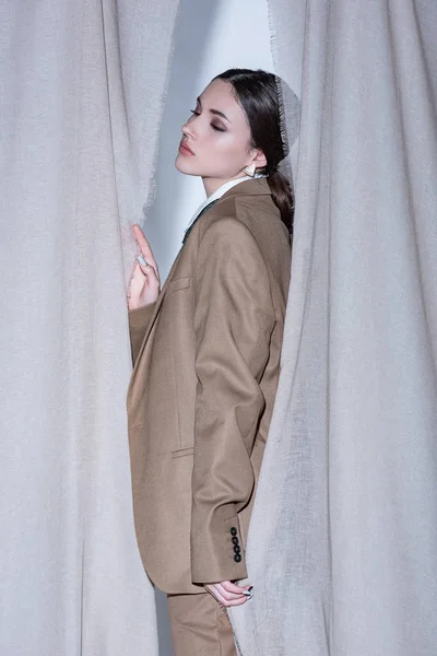 Vue latérale de la femme à la mode en costume debout sur fond de rideau gris clair — Photo de stock