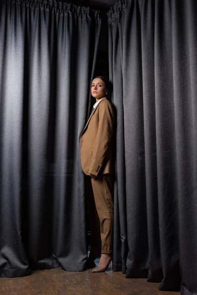 Вид збоку модна модель в костюмі, що стоїть на темно-сірому фоні штори, дивлячись на камеру — стокове фото