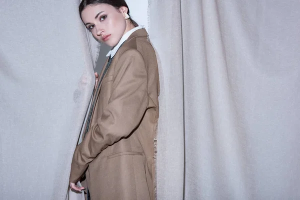 Side view of fashionable woman in suit standing on light grey curtain background, looking at camera — Stock Photo