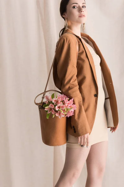 Fashionable woman with flowers in bag standing on curtain background, looking away — Stock Photo