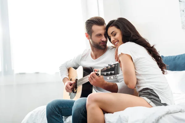 Mann spielt Akustikgitarre neben glücklicher Frau im Bett — Stockfoto