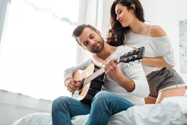 Vista basso angolo di uomo suonare la chitarra acustica vicino donna attraente a letto — Foto stock