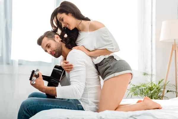 Attractive woman sitting on bed and hugging man with acoustic guitar — Stock Photo