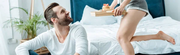 Panoramic shot of girlfriend giving tray with breakfast to smiling boyfriend — Stock Photo