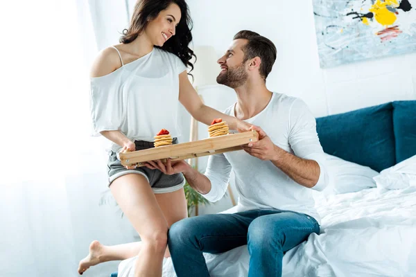 Happy girlfriend giving tray with breakfast to smiling boyfriend in bed — Stock Photo