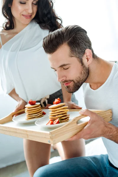 Joven oliendo sabrosos panqueques con fresas en bandeja de madera cerca de la novia sonriente - foto de stock