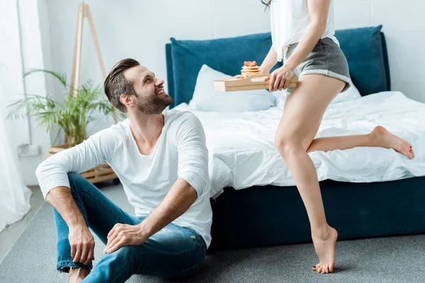 Vue recadrée de petite amie donnant plateau avec petit déjeuner à petit ami souriant — Photo de stock