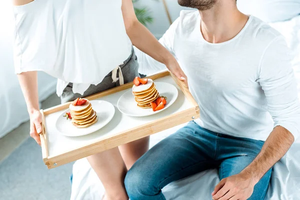 Partial view of woman holding wooden tray with delicious pancakes and strawberries on plates near man — Stock Photo