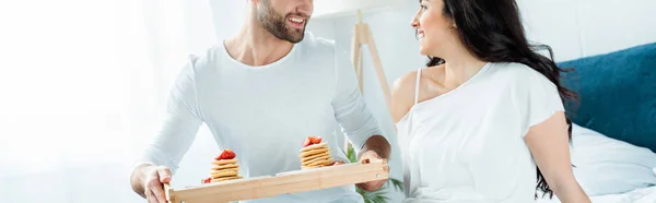 Panoramic shot of smiling man holding wooden tray with tasty pancakes near girlfriend — Stock Photo