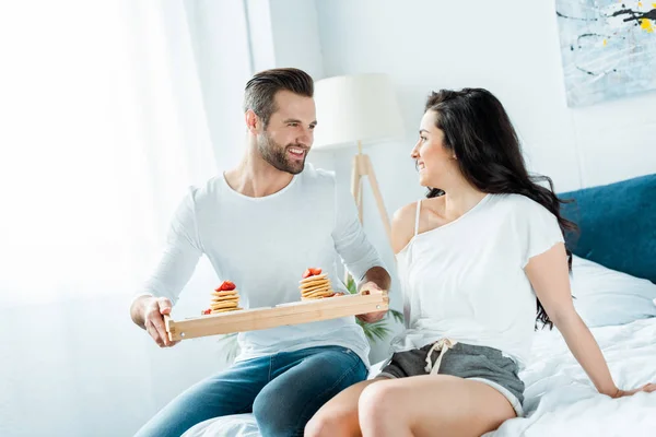 Lächelnder Freund mit Holztablett mit leckeren Pfannkuchen und Blick auf glückliche Freundin im Schlafzimmer — Stockfoto
